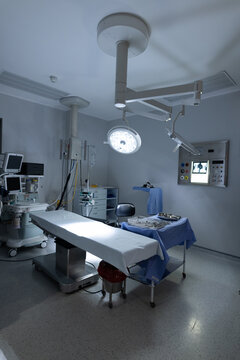Unoccupied operating room holding table with white sheet, blue drape, ceiling light and IV poles
