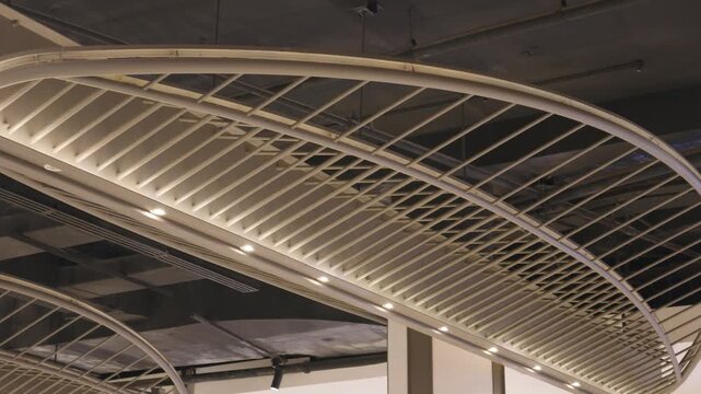 Close-up view of a modern industrial ceiling featuring curved metal beams, white linear ceiling slats, exposed concrete, ventilation ducts, and recessed lights.