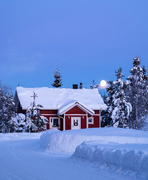 Cozy red cottage in a snow-covered pine forest under a fading moon at sunrise, scenic polar morning or Christmas vacation concept in Finland.