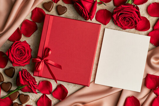 A high angle romantic composition featuring a red gift box with a satin bow a blank greeting card scattered rose petals lush red roses and heart shaped chocolates on a soft textured surface