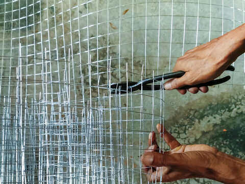 Worker hands cutting wire mesh with pliers on concrete floor. Hands using wire cutter pliers to cut roll of silver galvanized steel wire mesh fencing. DIY construction, craftsmanship, and manual labor