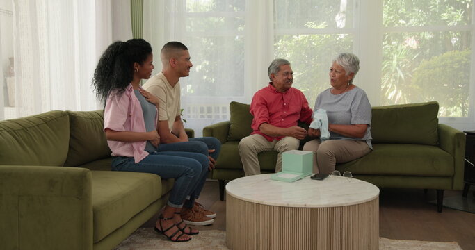 Diverse family gathering in living room, grandparents joyfully receiving baby clothes gift, at home