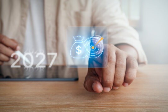 Businessman finger touching digital money bag and target icon overlay with year 2027 text, concept of financial target, business strategy planning, and budget goal for 2027.