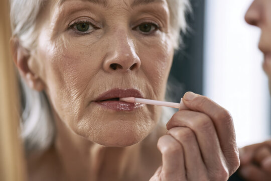 A graceful older woman delicately applies lip gloss with a keen focus in front of a mirror, showcasing beauty, self-care, and confidence in her daily routine.