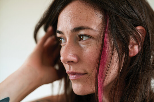 A close-up image of a contemplative woman with long hair and a striking pink highlight. Her expression captures a moment of introspection and beauty.