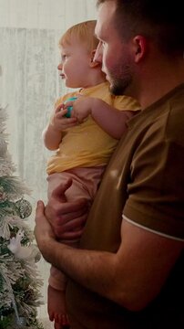 A Heartwarming Moment: Father and Toddler Decorate the Christmas Tree Together, Creating Lasting Memories in a Cozy Holiday Setting