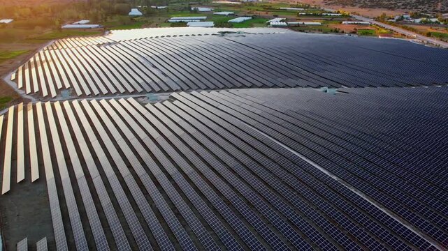 Aerial view of a vast solar power plant with long rows of photovoltaic panels reflecting warm sunlight. Rural fields and farm buildings surround the large renewable energy facility in the countryside.