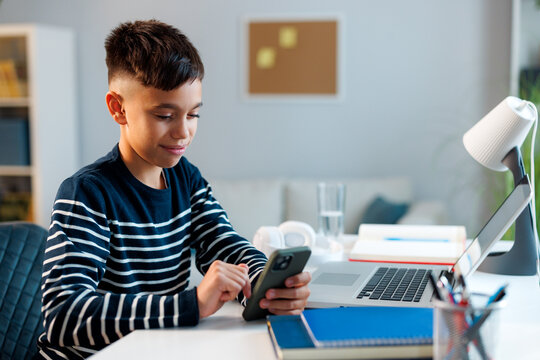 Boy tapping a smartphone at his desk beside an open laptop, combining digital tools for schoolwork, online research, and independent learning in a home study space