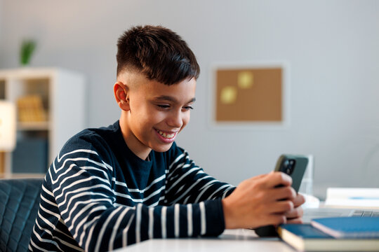 Boy smiling and holding a smartphone, engaging with digital communication and online learning at a desk in a bright home study area with books nearby