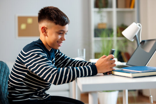 Boy smiling while checking a smartphone beside an open laptop and school books, combining digital communication with home learning at a bright study desk