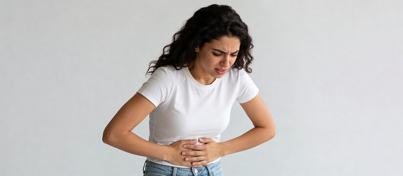 Woman clutching abdomen while looking down against plain gray background. Abdominal pain and stomachache symptom. Digestive health and medical care concept. Illness and healthcare awareness.