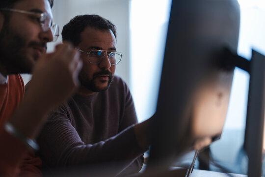 Two male colleagues wearing glasses collaborate in a modern office. They sit together at a desk, looking closely at a computer monitor and discussing a project.