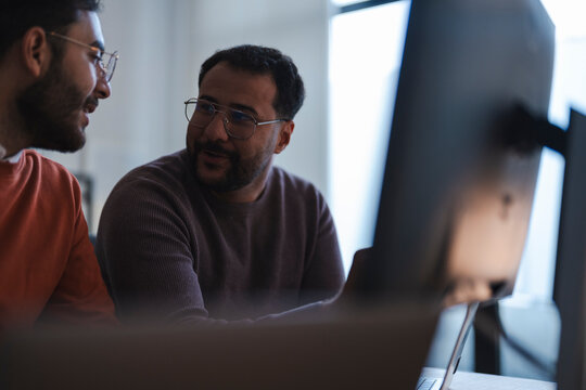 Two male colleagues are sitting together in a modern office. They are talking and collaborating on a project while looking at a computer screen during the workday.