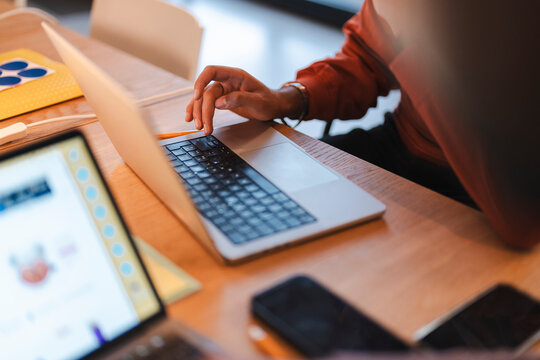 An office worker wearing a red sweater sits at a wooden desk and types on a laptop keyboard during a busy workday inside a bright contemporary office space.