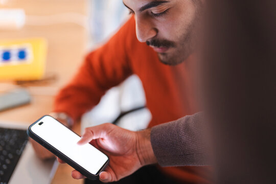 A young man wearing a red sweater sits at a desk while holding a modern smartphone. He taps the bright, blank screen with his finger, focusing on the mobile device.