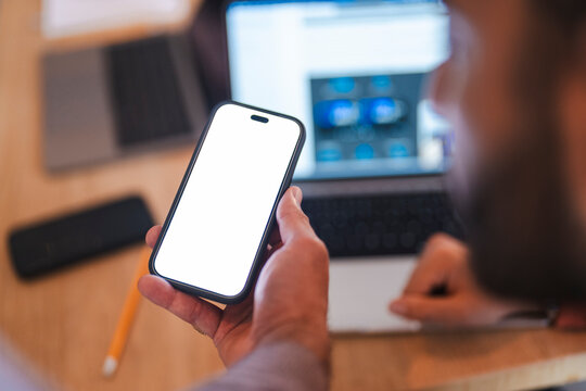 A man holding a modern smartphone with a blank white screen mockup. He sits at a wooden desk with a laptop in the blurred background, working in an office environment.