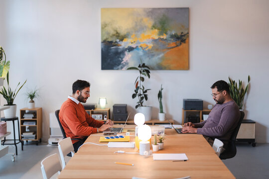 Two male colleagues sit at a long wooden table in a bright modern workspace. They are focused on their individual laptops, typing and working on business projects.