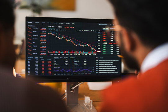 A financial trader analyzes stock market charts and data on a computer screen, pointing at a decreasing currency graph while working in a modern home office.