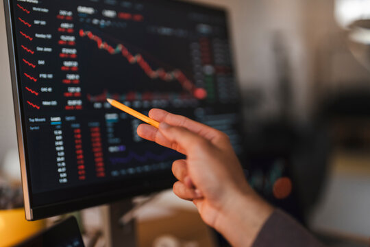 A financial analyst points a pencil at a computer screen showing stock market charts with declining red trends. The person evaluates economic data in an office.