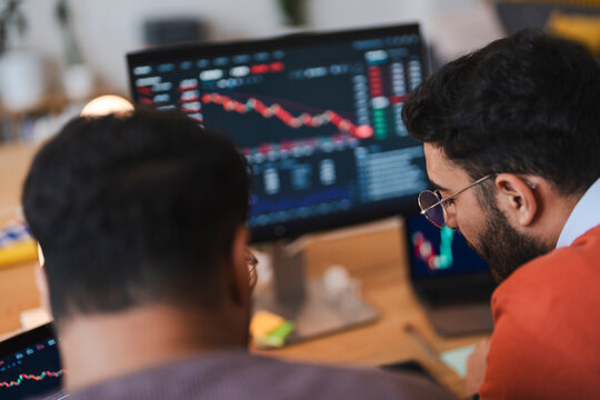 Two male financial traders analyze stock market data and financial charts on multiple computer screens inside a modern office workspace during evening hours.