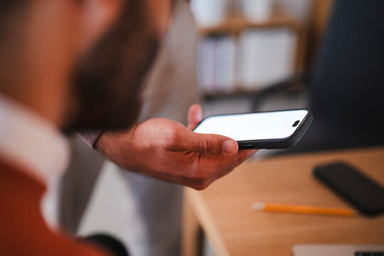 A bearded man holding a smartphone with a blank white screen horizontally inside a bright office. He is looking at the device display during a professional workplace meeting.
