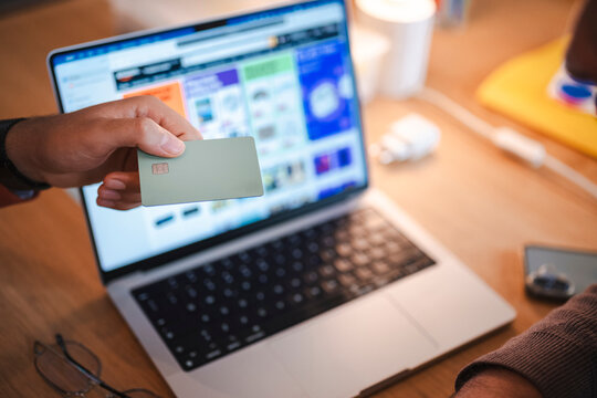 A person holds a green payment card in front of a laptop computer. They are sitting at a wooden desk, getting ready to make an online purchase using their credit card.