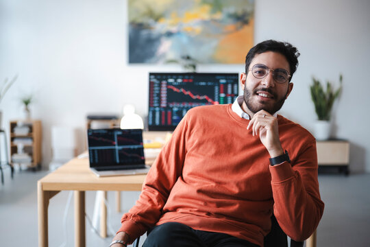 A smiling young man with glasses sits at a desk in an office. Computer monitors in the background display financial charts and trading data while he works remotely.