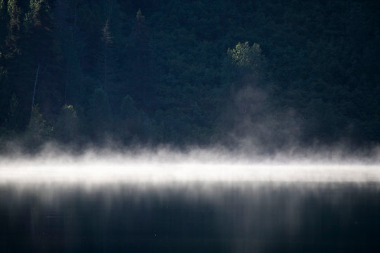 Fog over the water with green trees on a hill in the background at a lake near Soldotna, Alaska on a summer day.