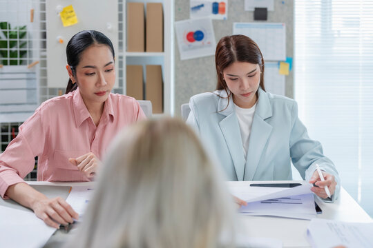 Two serious Asian businesswomen reviewing resume and documents while interviewing job candidate in office, HR recruitment and hiring assessment concept 