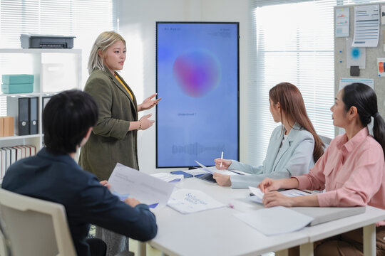 Asian business team listening to colleague presenting project update in front of screen and bulletin board with sticky notes, charts and schedule in office