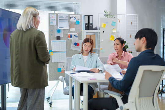 Asian business team listening to colleague presenting project update in front of screen and bulletin board with sticky notes, charts and schedule in office