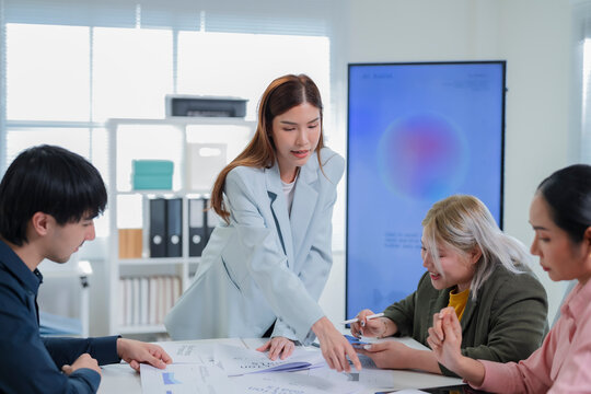 Asian businesswoman leaning over table and pointing at business proposal while guiding team through quarterly report in modern office with AI assistant screen