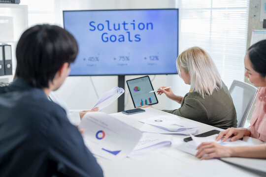Business team reviewing solution goals and growth targets on screen while colleague points at charts on digital tablet during strategy meeting in office