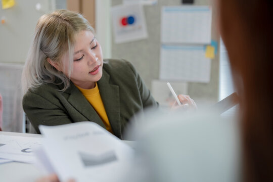 Focused young Asian businesswoman with dyed blonde hair taking notes with stylus on digital tablet while reviewing documents during office meeting