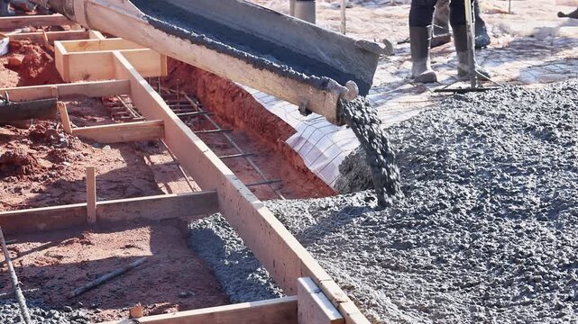 Wet concrete pours from chute into rectangular foundation frame on red soil ground