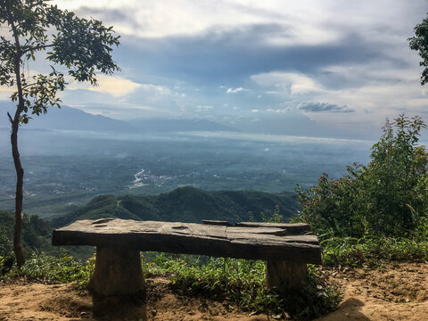 A rustic wooden bench placed on a viewpoint cliff overlooking lush green mountains, sea of mist, and cloudy sky in the morning, perfect for travel, nature, and relaxation concepts.