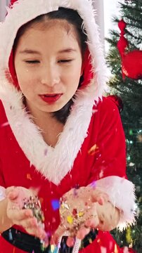 Girl in Santa costume blows into cupped hands by Christmas tree