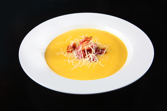 Creamy yellow soup in white bowl. Crispy bacon and grated cheese garnish. Black background highlights dish. Gourmet appetizer