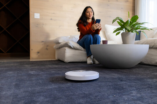 Asian woman sitting on sofa holding smartphone near robotic vacuum on dark carpet