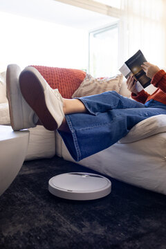 Woman in red-orange sweater reclining on sofa reading open book near white robot vacuum on rug