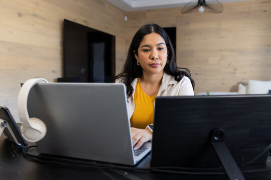 Woman with long dark hair typing on silver laptop beside second display in home office