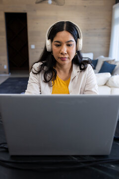 Woman wearing light jacket and headphones sitting at dark table facing open laptop in living room