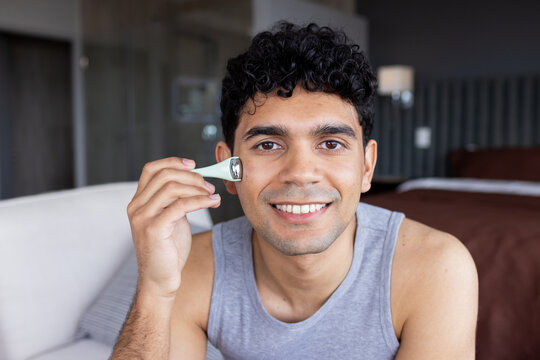 African American man holding pale green facial roller against right cheek while smiling in room