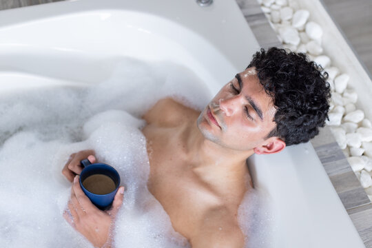 African American man reclining in white bathtub holding blue mug above thick foam while relaxing