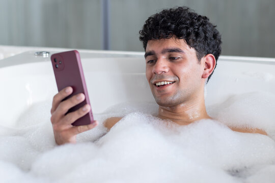 African American man sitting in bathtub filled with thick foam while gazing at smartphone