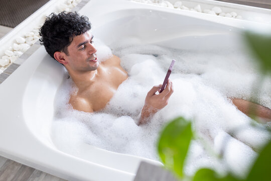 African American man holding smartphone while reclining in white bathtub with thick foam