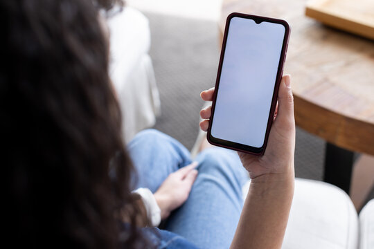 Wavy-haired person sitting while holding smartphone with blank screen near round table, light sofa