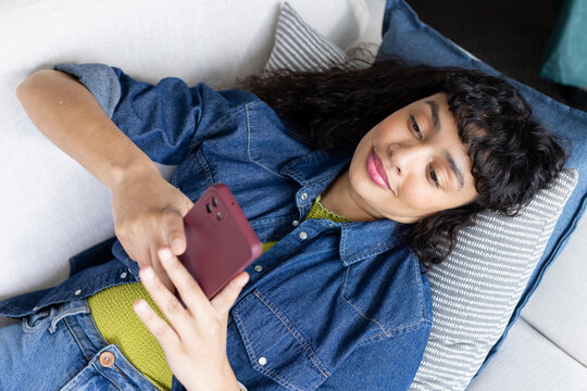 African American woman reclining on sofa holding burgundy smartphone and looking at screen