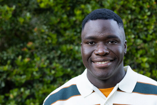African American man in striped collared shirt smiling at camera beside green hedge