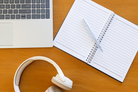 White laptop sitting on wooden desk left beside spiral notebook holding white pen and headphones